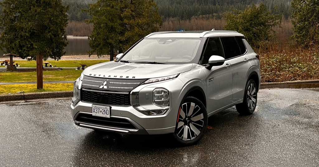 2026-Outlander-PHEV - Portage La Prairie Voice Front-three-quarter view of a silver Mitsubishi Outlander SUV parked on a wet pavement with trees and a lake in the background, daytime outdoor scene.